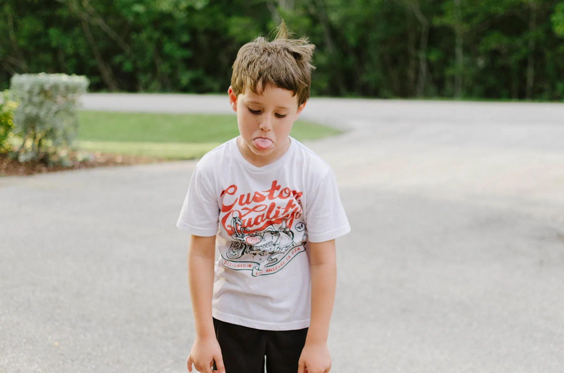 Young boy sticking out his tongue in frustration while standing outdoors on a quiet suburban street.