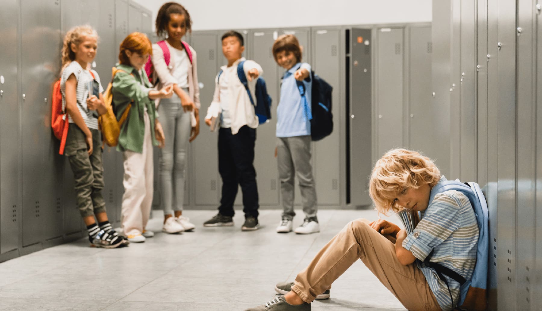 Sad crying schoolboy pupil sitting on the floor at the school hall while his classmates teenagers bully and tease him