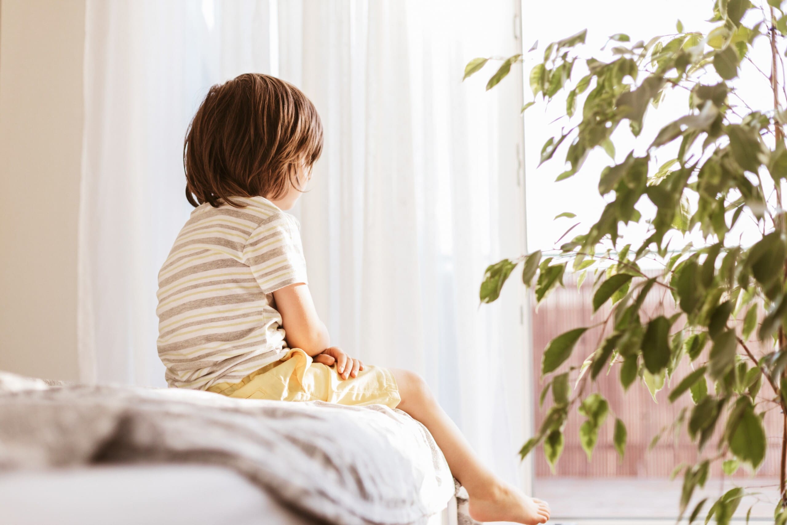 Child sitting on a bed looking out the window. Recognizing children's mental health.