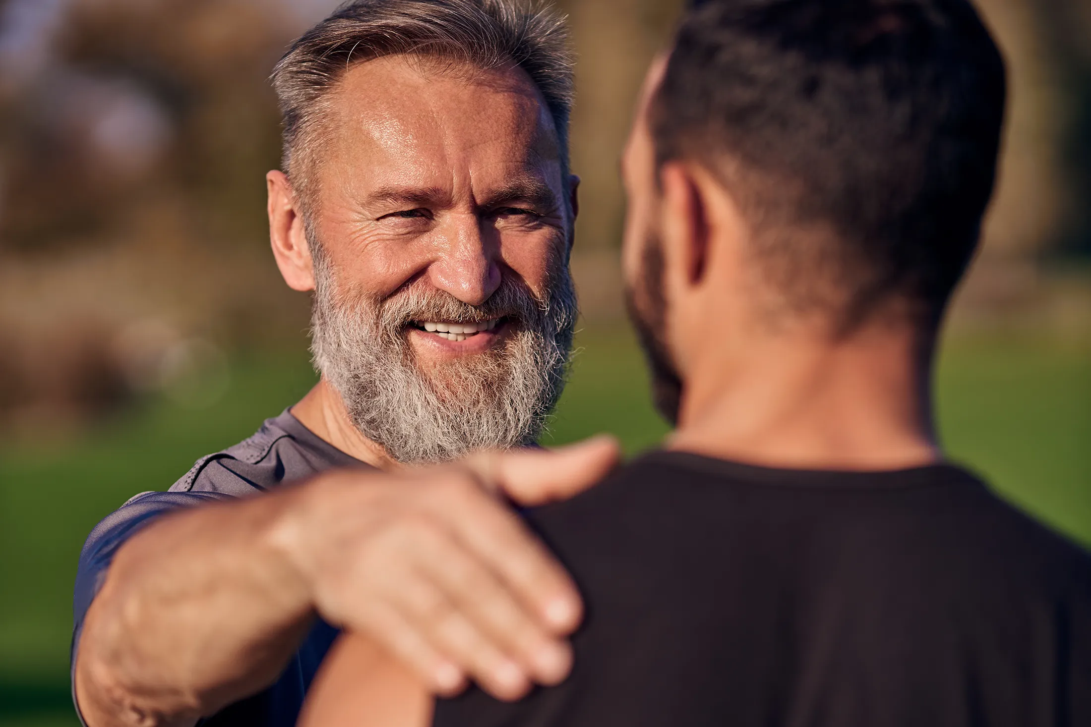 Smiling older man placing a supportive hand on a younger man's shoulder outdoors.