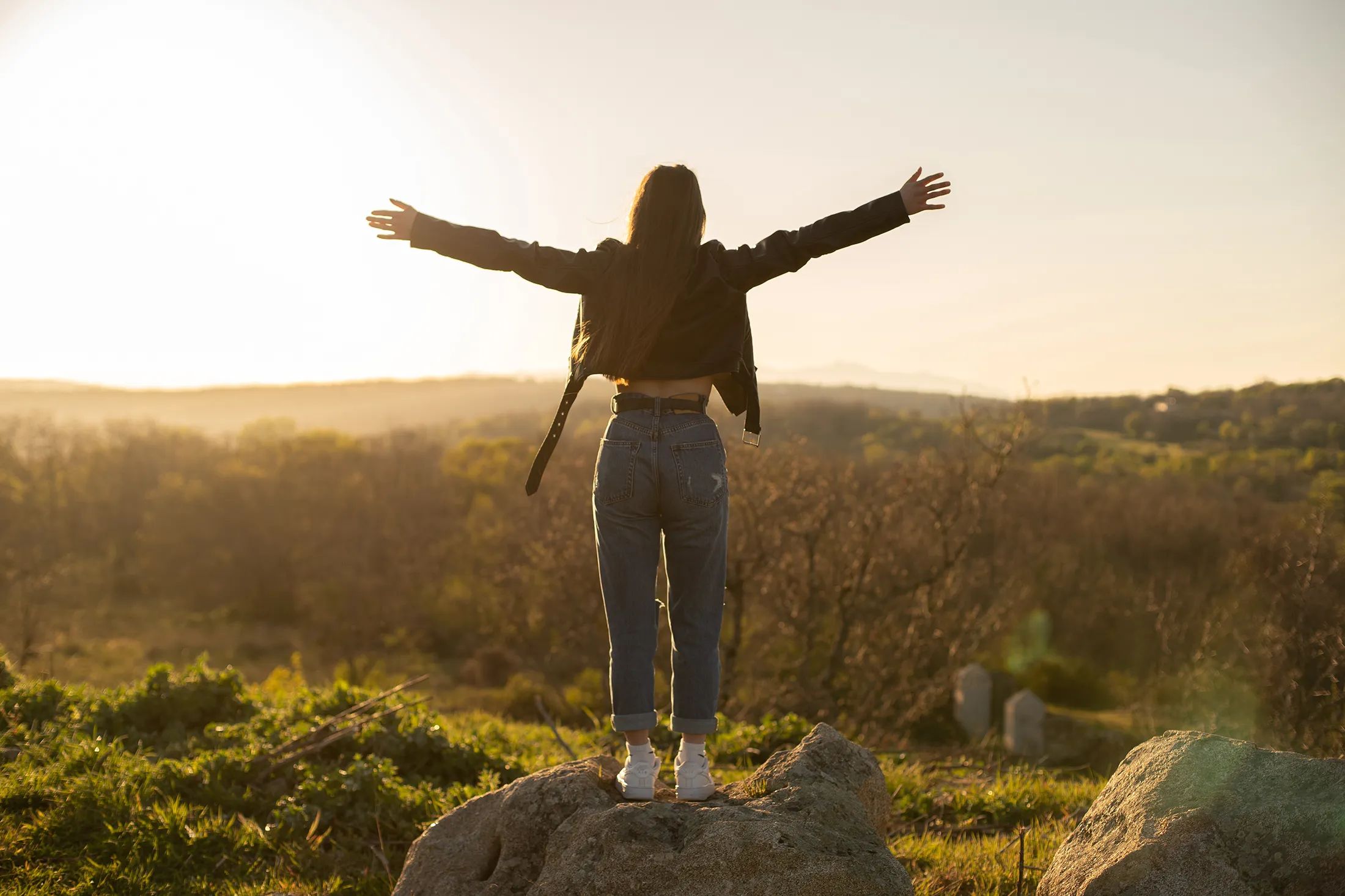 Woman standing on a rock with arms outstretched, embracing the sunset over a scenic field.